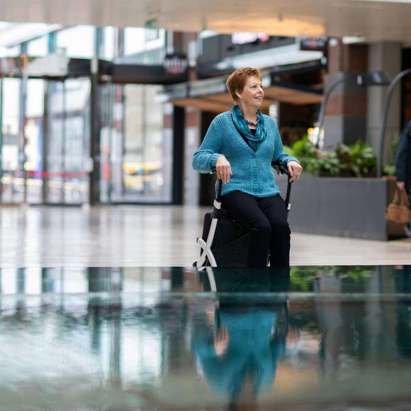 Senior souriante assise sur siège intégré du rollator Rollz Flex blanc dans un centre commercial