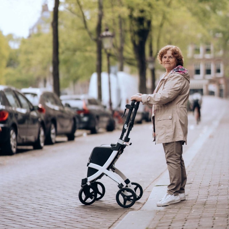 Senior debout dans une rue pavée tenant le rollator Rollz Flex blanc replié et compact
